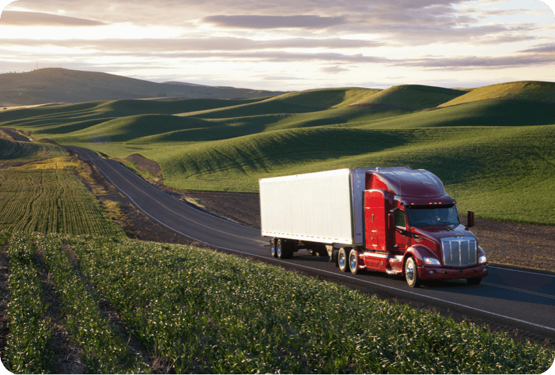 Red and white semi truck driving down a black asphalt road with grass and hills on both sides of the road